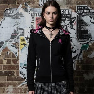 May include: Black zip-up hoodie with a pink skull design on the chest and hood lining. The model is wearing a black choker, a plaid skirt, and ripped black jeans. The background is a brick wall with torn posters.