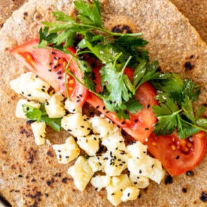 May include: A close-up shot of a wrap filled with fresh ingredients. The wrap is filled with sliced red tomatoes, white cheese, and green parsley. Black sesame seeds are sprinkled on the tomatoes and cheese. The wrap is a light brown color.