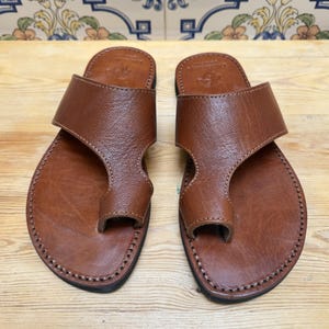 May include: A pair of brown leather sandals with a toe loop and a wide strap across the top. The sandals are stitched around the edges and rest on a light brown wooden surface. The background features decorative tiles.