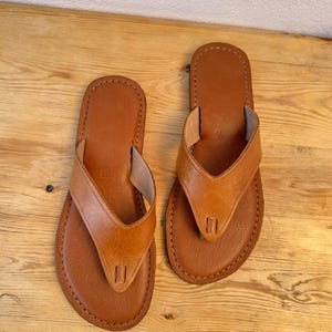 May include: A pair of brown leather flip-flops with a stitched design. The sandals have a thong strap and a rounded toe. The leather appears to be a light brown color. The sandals are displayed on a wooden surface.