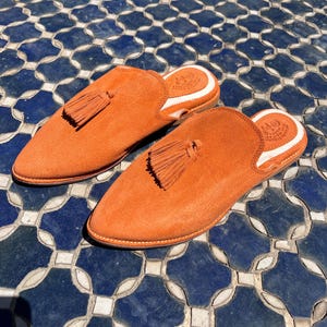May include: A pair of orange suede slip-on shoes with pointed toes and a tassel detail. The shoes have a white trim around the opening and are set on a blue and white tiled surface. The shoes are a vibrant shade of orange.