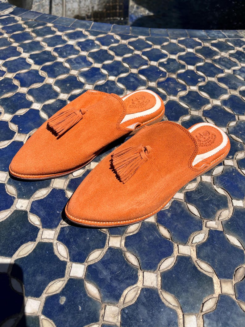 May include: A pair of orange suede slip-on shoes with pointed toes and a tassel detail. The shoes have a white trim around the opening and are set on a blue and white tiled surface. The shoes are a vibrant shade of orange.