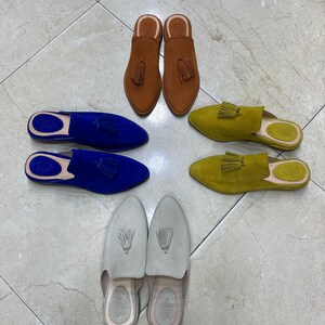 May include: An assortment of suede slip-on shoes in various colors, including brown, blue, yellow-green, and light gray. Each shoe features a pointed toe and a tassel detail. The shoes are arranged on a tiled surface.