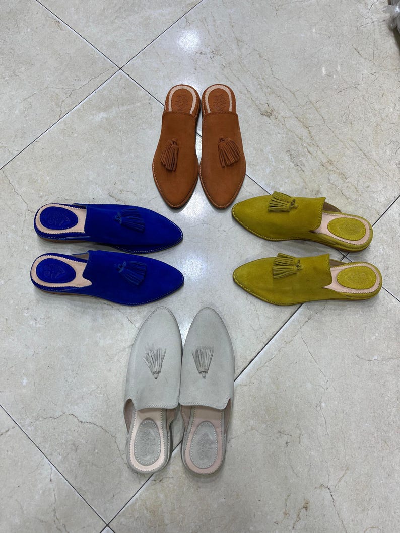 May include: An assortment of suede slip-on shoes in various colors, including brown, blue, yellow-green, and light gray. Each shoe features a pointed toe and a tassel detail. The shoes are arranged on a tiled surface.