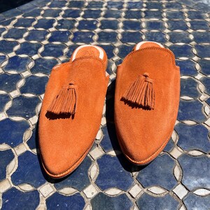 May include: A pair of orange suede slip-on shoes with a tassel detail. The shoes have a rounded toe and a white interior lining. The shoes are displayed on a blue and white tiled surface.