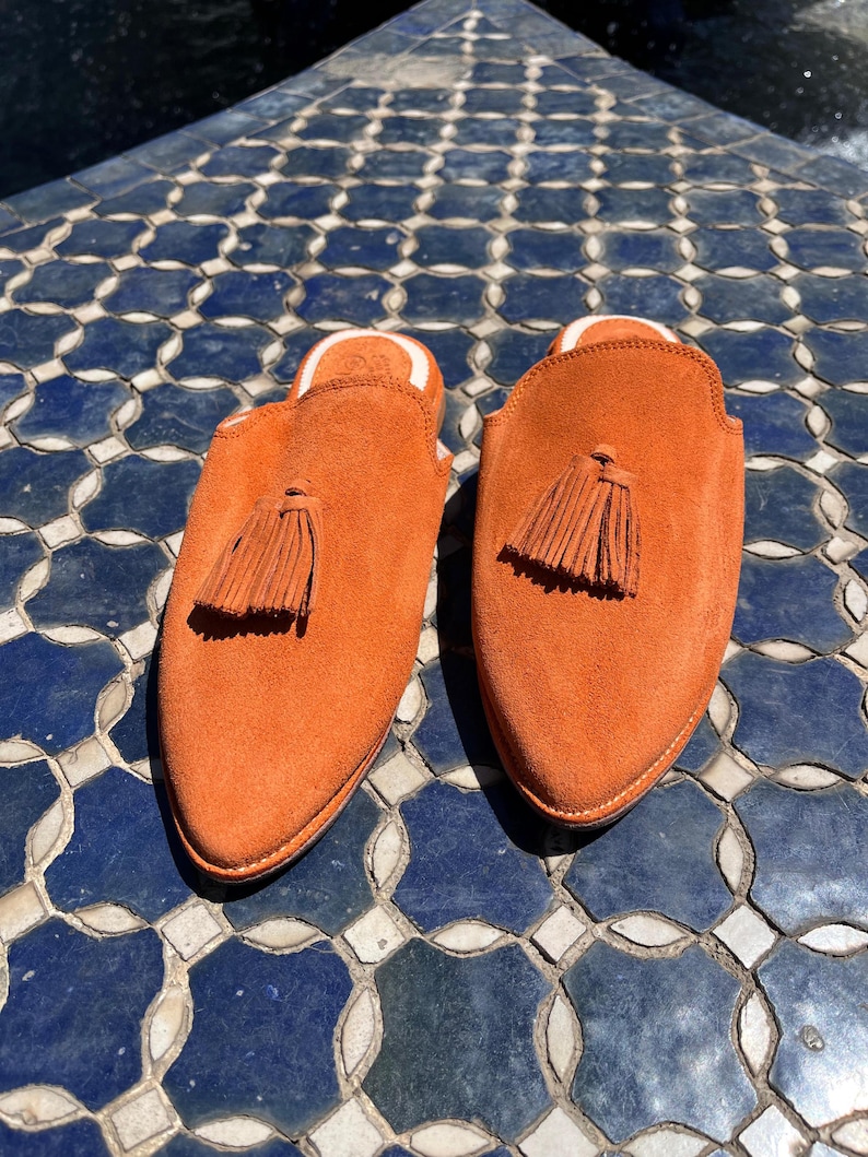 May include: A pair of orange suede slip-on shoes with a tassel detail. The shoes have a rounded toe and a white interior lining. The shoes are displayed on a blue and white tiled surface.