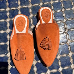 May include: A pair of orange suede slip-on shoes with a pointed toe and a small tassel. The shoes have a leather insole with a logo and a white trim. The shoes are displayed on a blue and white tile surface.