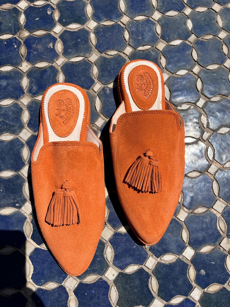 May include: A pair of orange suede slip-on shoes with a pointed toe and a small tassel. The shoes have a leather insole with a logo and a white trim. The shoes are displayed on a blue and white tile surface.
