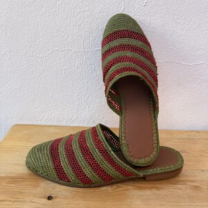 May include: A pair of woven slippers with a green and red striped pattern. The slippers have a brown leather insole and a flat sole. The design is open-backed, and the slippers are displayed on a wooden surface.