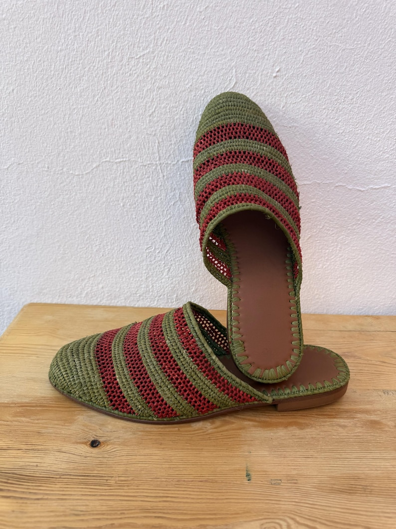May include: A pair of woven slippers with a green and red striped pattern. The slippers have a brown leather insole and a flat sole. The design is open-backed, and the slippers are displayed on a wooden surface.