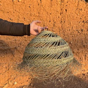 May include: A woven basket with a conical shape, crafted from light green and tan natural fibers. The basket has an open design with a textured surface and a fringe of loose fibers at the bottom. The background is a textured, reddish-brown wall.