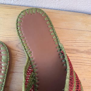 May include: Close-up of a woven sandal with a brown insole and green stitching. The sandal's upper features alternating red and green stripes, creating a textured appearance. The design suggests a focus on comfort and style.