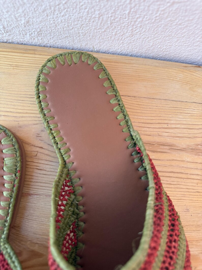 May include: Close-up of a woven sandal with a brown insole and green stitching. The sandal's upper features alternating red and green stripes, creating a textured appearance. The design suggests a focus on comfort and style.
