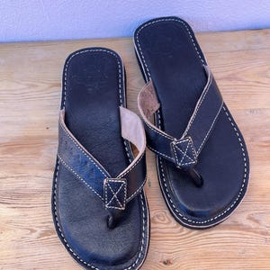 Könnte beinhalten: Ein Paar schwarze Leder-Flip-Flops mit weißen Nähten. Die Sandalen haben einen Zehenriemen und ein genähtes X-Detail. Die Sohlen sind dunkel und die Sandalen werden auf einer Holzoberfläche präsentiert.