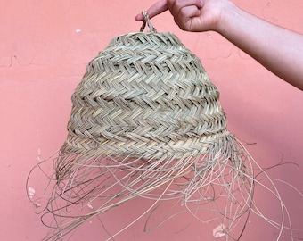 Puede incluir: Una cesta en forma de campana, tejida a mano, de color marrón claro natural. La cesta presenta un patrón tejido y largos mechones colgantes en la parte inferior. Se sostiene sobre un fondo rosa.