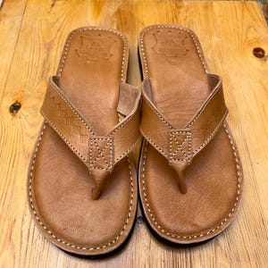 Pode incluir: Um par de chinelos de couro castanho com design de tira de dedo. As sandálias têm uma borda costurada e um detalhe de tira texturizada. As sandálias são exibidas em uma superfície de madeira.