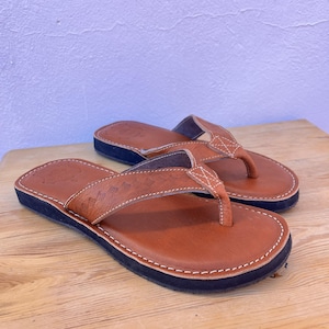 Puede incluir: Un par de chanclas de cuero marrón con suela azul oscuro y costuras blancas. Las sandalias tienen un diseño de correa de tanga y se exhiben sobre una superficie de madera.