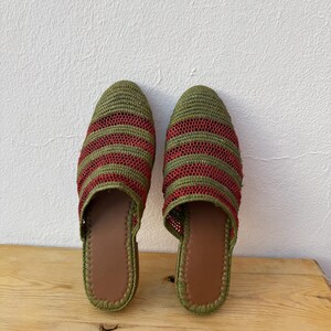 May include: A pair of handmade green and red striped woven slippers. The slippers have a brown leather insole and a green trim. The slippers are displayed on a wooden surface.