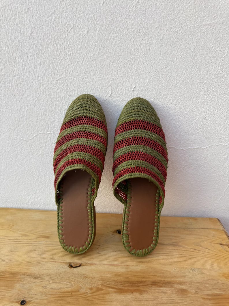 May include: A pair of handmade green and red striped woven slippers. The slippers have a brown leather insole and a green trim. The slippers are displayed on a wooden surface.