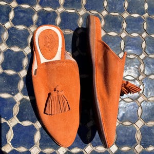 May include: A pair of orange suede slip-on slippers with a tassel detail. The slippers have a rounded toe and a low heel. The interior is a light tan color. The slippers are displayed on a blue and white tiled surface.