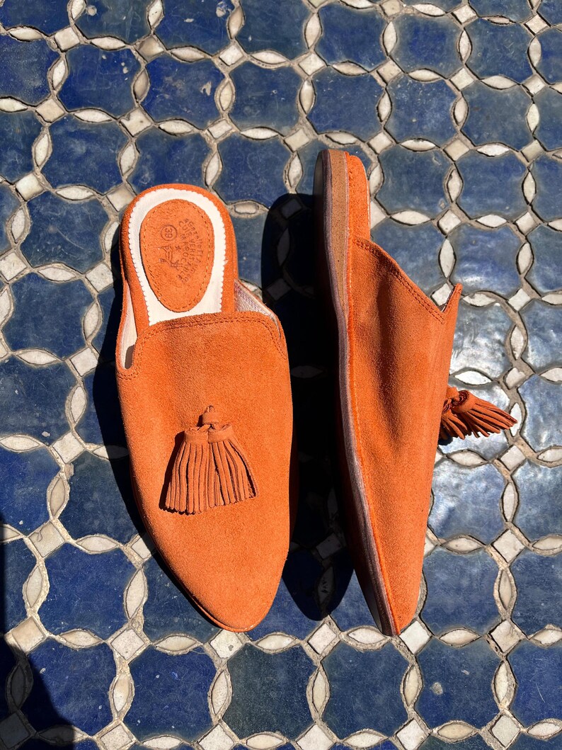 May include: A pair of orange suede slip-on slippers with a tassel detail. The slippers have a rounded toe and a low heel. The interior is a light tan color. The slippers are displayed on a blue and white tiled surface.