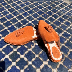 May include: A pair of orange suede slip-on shoes with tassel details. The shoes have a rounded toe and a white interior. The shoes are displayed on a blue and white tile surface.