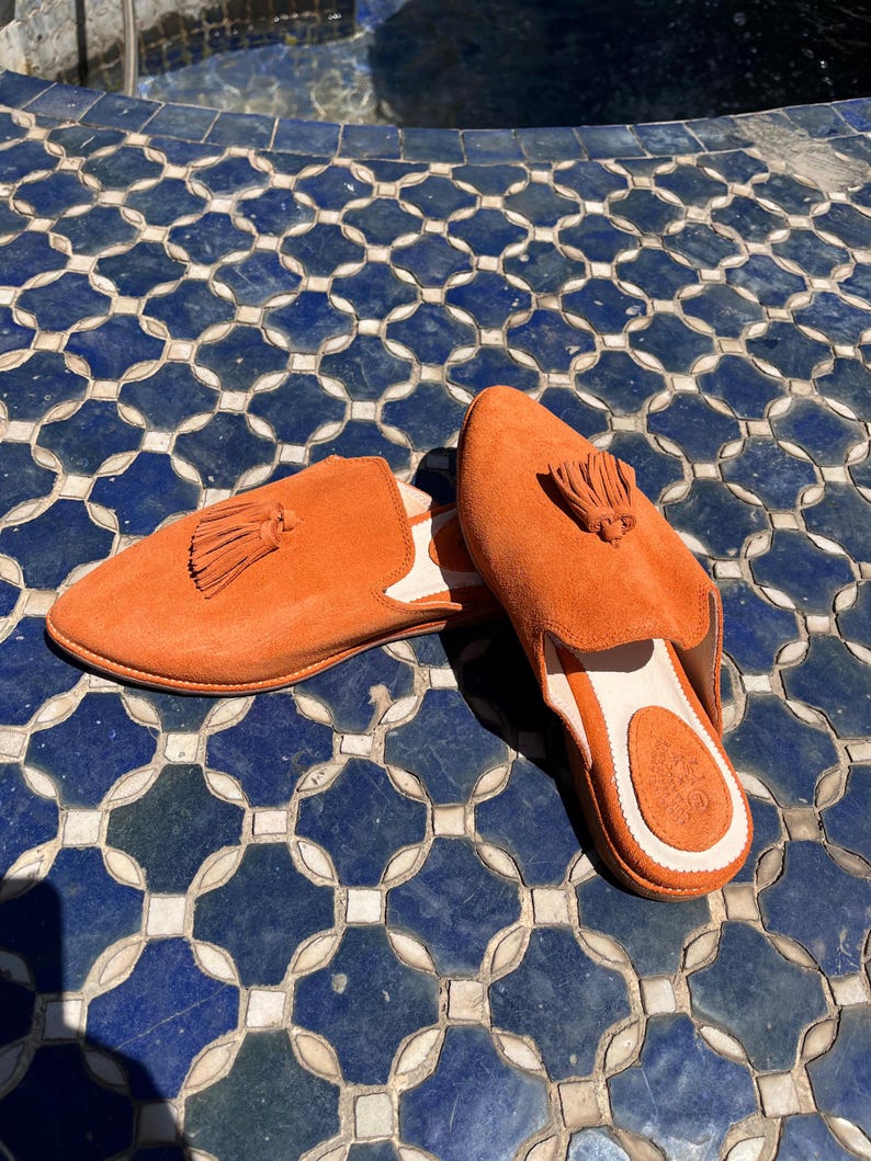 May include: A pair of orange suede slip-on shoes with tassel details. The shoes have a rounded toe and a white interior. The shoes are displayed on a blue and white tile surface.