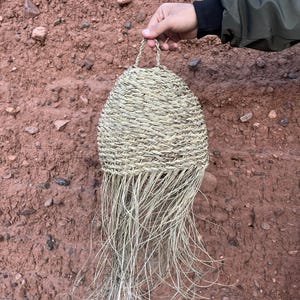 May include: A hand-woven basket with a rounded body and a looped handle. The basket is made of natural-colored woven material, with long, loose strands extending from the bottom. The basket is held by a person.