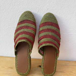 May include: A pair of handmade green and red striped woven slippers. The slippers have a brown leather insole and a green stitched border. The slippers are displayed on a wooden surface against a white wall.
