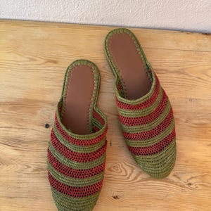 May include: A pair of handmade green and red striped woven slippers. The slippers have a brown leather insole and a rounded toe. The slippers are displayed on a light brown wooden surface.