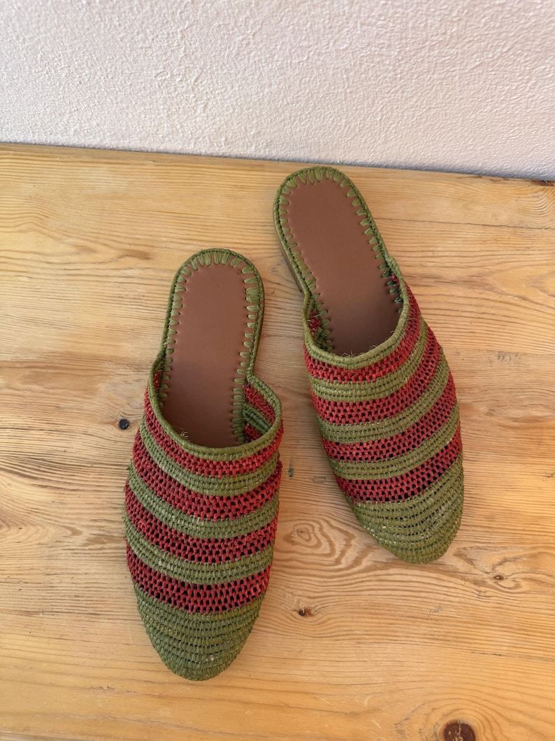 May include: A pair of handmade green and red striped woven slippers. The slippers have a brown leather insole and a rounded toe. The slippers are displayed on a light brown wooden surface.