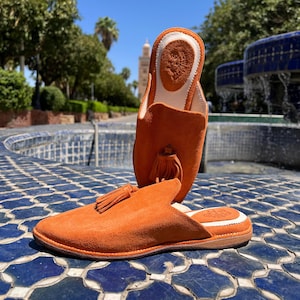 May include: A pair of orange suede slippers with a tassel detail. The slippers are open-backed and have a leather sole. One slipper is upright, showcasing the interior with a logo. The background features a blue tiled surface and a fountain.