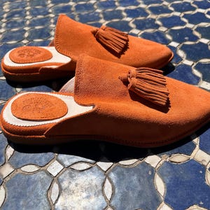 May include: A pair of orange suede slippers with a tassel detail. The slippers have a rounded toe and a white and brown leather insole. The slippers are displayed on a blue and white tile surface.