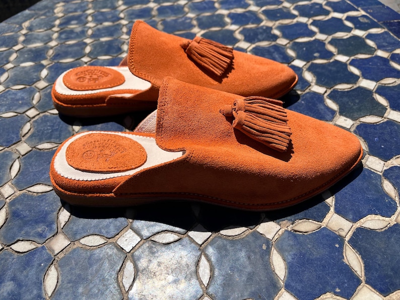May include: A pair of orange suede slippers with a tassel detail. The slippers have a rounded toe and a white and brown leather insole. The slippers are displayed on a blue and white tile surface.