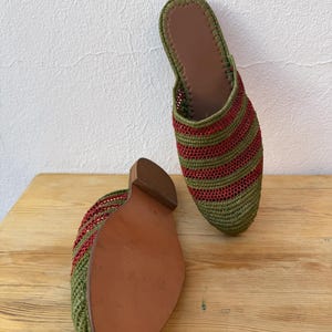 May include: A pair of green and red striped woven slippers with brown leather soles. The slippers are displayed on a wooden surface, showcasing their design and construction. The open-back style and color scheme suggest a casual, comfortable footwear option.