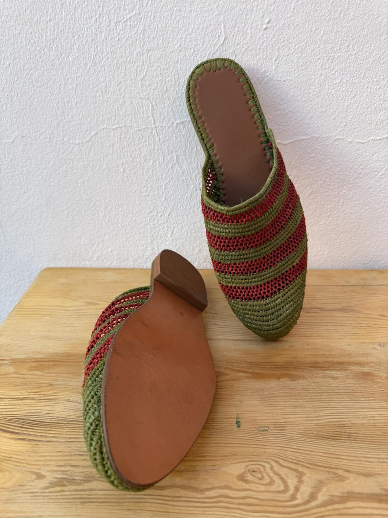 May include: A pair of green and red striped woven slippers with brown leather soles. The slippers are displayed on a wooden surface, showcasing their design and construction. The open-back style and color scheme suggest a casual, comfortable footwear option.