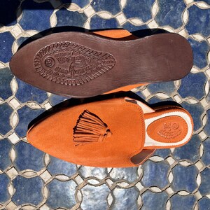 May include: Orange suede slip-on shoes with a tassel detail. The shoes have a brown leather sole with embossed details. The interior is white with a stamped logo. The shoes are displayed on a blue and white tile surface.