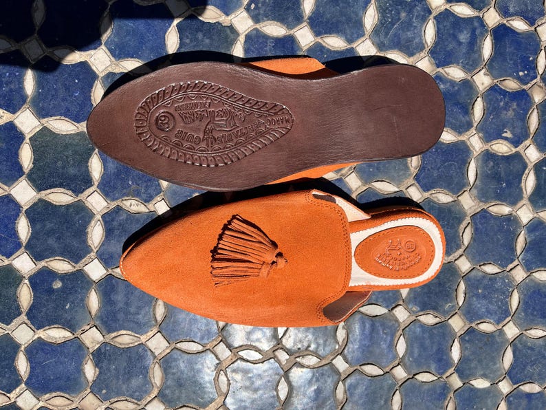 May include: Orange suede slip-on shoes with a tassel detail. The shoes have a brown leather sole with embossed details. The interior is white with a stamped logo. The shoes are displayed on a blue and white tile surface.