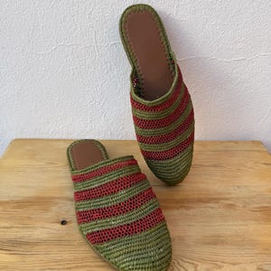 May include: A pair of woven raffia slippers. The slippers are green with horizontal red stripes. The interior is brown. The slippers are displayed on a wooden surface against a white wall.