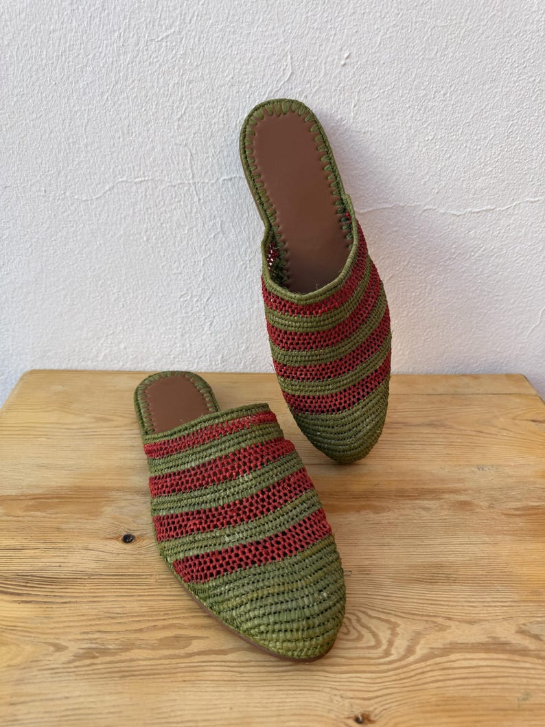 May include: A pair of woven raffia slippers. The slippers are green with horizontal red stripes. The interior is brown. The slippers are displayed on a wooden surface against a white wall.