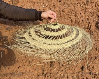 May include: A woven, natural fiber lampshade with a circular design. The lampshade is made of light beige material and has a layered, spiral pattern. The outer edges have long, flowing strands, creating a fringed effect. The lampshade is held by a hand.