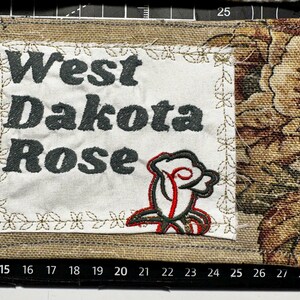 May include: Embroidered patch featuring the text "West Dakota Rose" in dark grey. A red, white, and green rose emblem is below the text. The patch is bordered with a floral design and a burlap-like fabric.