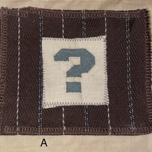 May include: A rectangular fabric patch with a brown striped background and a white square in the center. A blue question mark is stitched onto the white square. The patch has a stitched border and the letter "A" is visible.