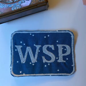 Puede incluir: Un parche rectangular de tela con fondo azul oscuro que presenta un patrón de estrellas blancas y las letras WSP bordadas en blanco en el centro. El parche tiene un borde cosido en color blanco y esquinas redondeadas.