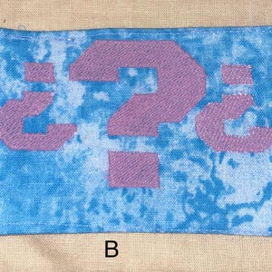 May include: A rectangular patch with a blue tie-dye background and a large, embroidered purple question mark. The letter "B" is at the bottom of the patch.