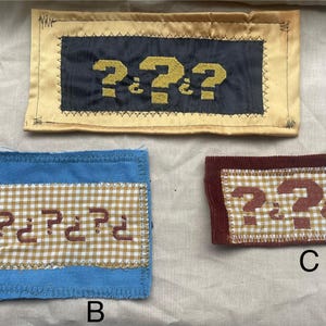 May include: Three fabric patches with question marks. The top patch has a black background with yellow question marks and a gold border. The bottom left patch is blue with a gingham pattern and red question marks. The bottom right patch is burgundy with a gingham pattern and red question marks.