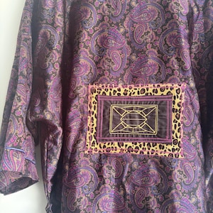 May include: A long-sleeved garment featuring a dark purple and black paisley pattern. A rectangular patch is centered on the back, consisting of a leopard print border surrounding a dark brown center with a yellow geometric embroidered design.