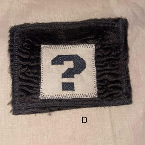 May include: A rectangular patch with a dark, textured border and a white square in the center. The white square features a navy blue question mark. The letter "D" is visible at the bottom.
