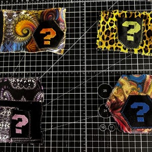May include: Four fabric patches with a black question mark on a black background. The patches have different patterns: a swirling design, leopard print, paisley, and a colorful abstract pattern. The patches are stitched onto a black grid.