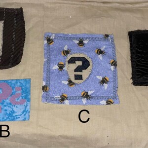 May include: Four fabric patches with question mark designs. One patch is brown with a white square and a blue question mark. Another is blue with a pink question mark. A third is blue with bee patterns and a black question mark. The last is black with a white square and a blue question mark.